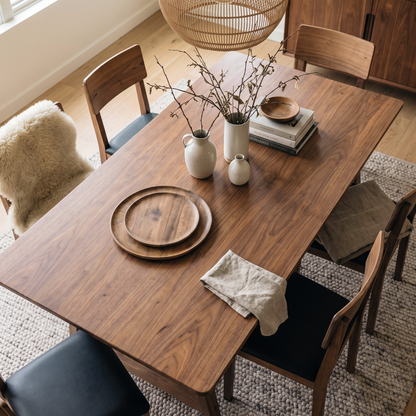 The Lucia Dining Table in warm walnut wood, styled in a minimalist dining room with chairs, a rug, and decorative accents.